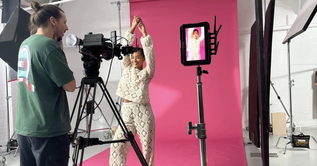 A model being filmed on a pink background with a camera operator stood behind camera.