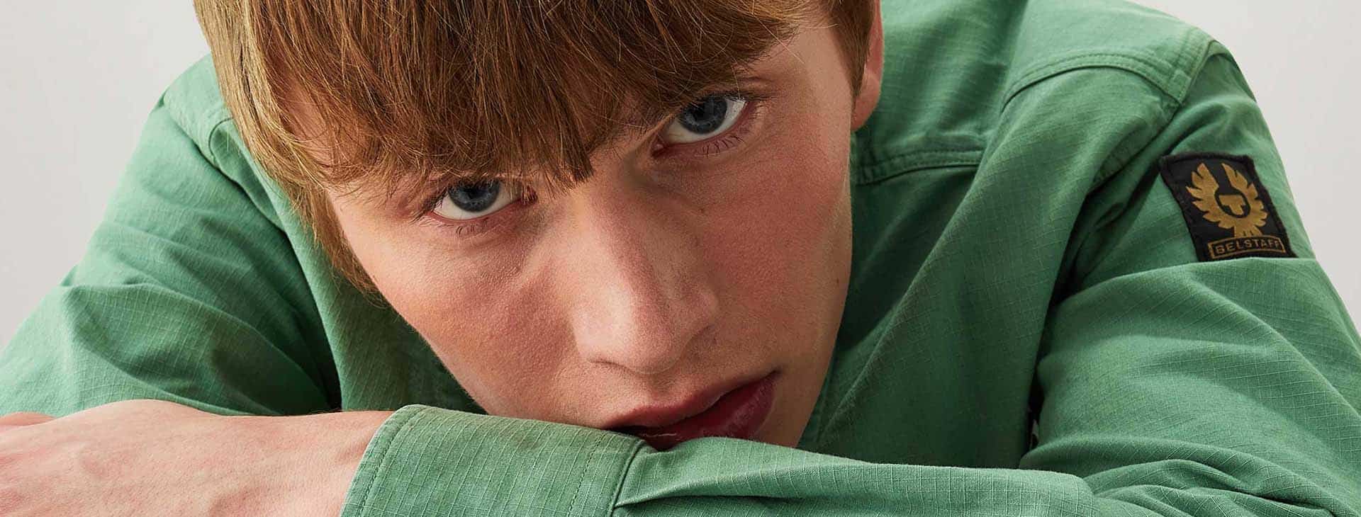 Man wearing green Belstaff shirt leaning on a table looking at camera.