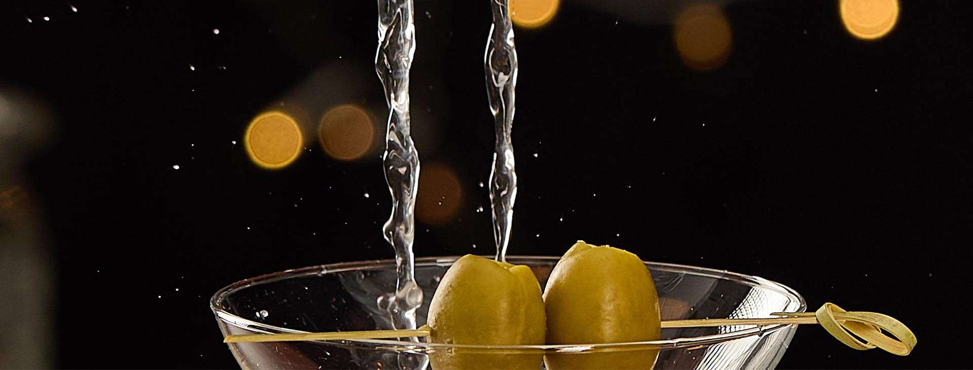 The rim of a martini glass with two olives on a stick and liquid being poured in.