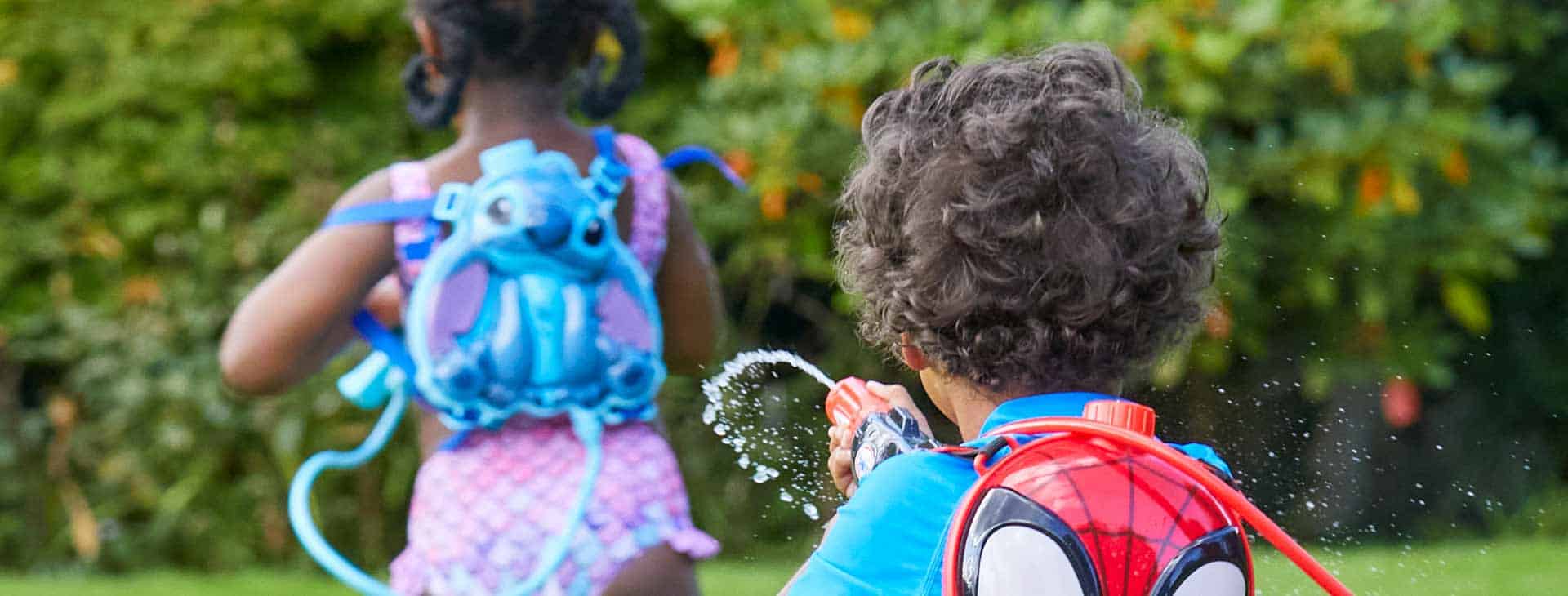 Two children playing with water pistols in garden wearing Disney character backpacks.